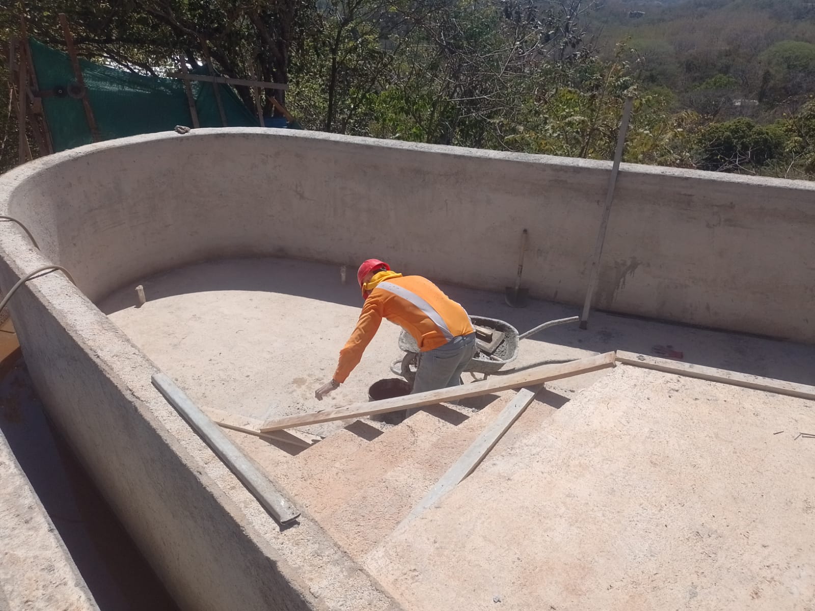 Un trabajador con casco rojo y camisa naranja realiza labores de construcción en una piscina en obra gris.