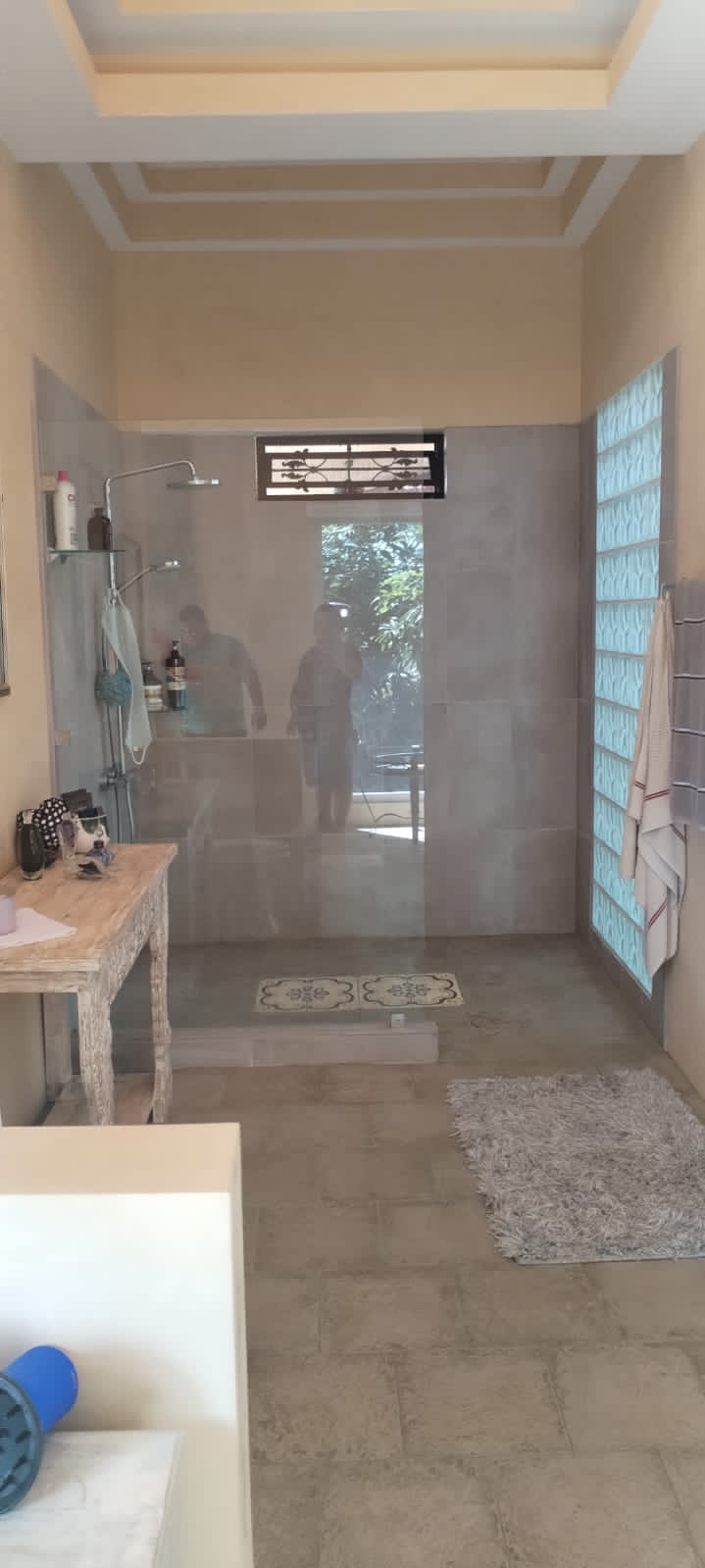 Se muestra el interior de un baño con una ducha de cristal, paredes de azulejos y una mesa de madera rústica.