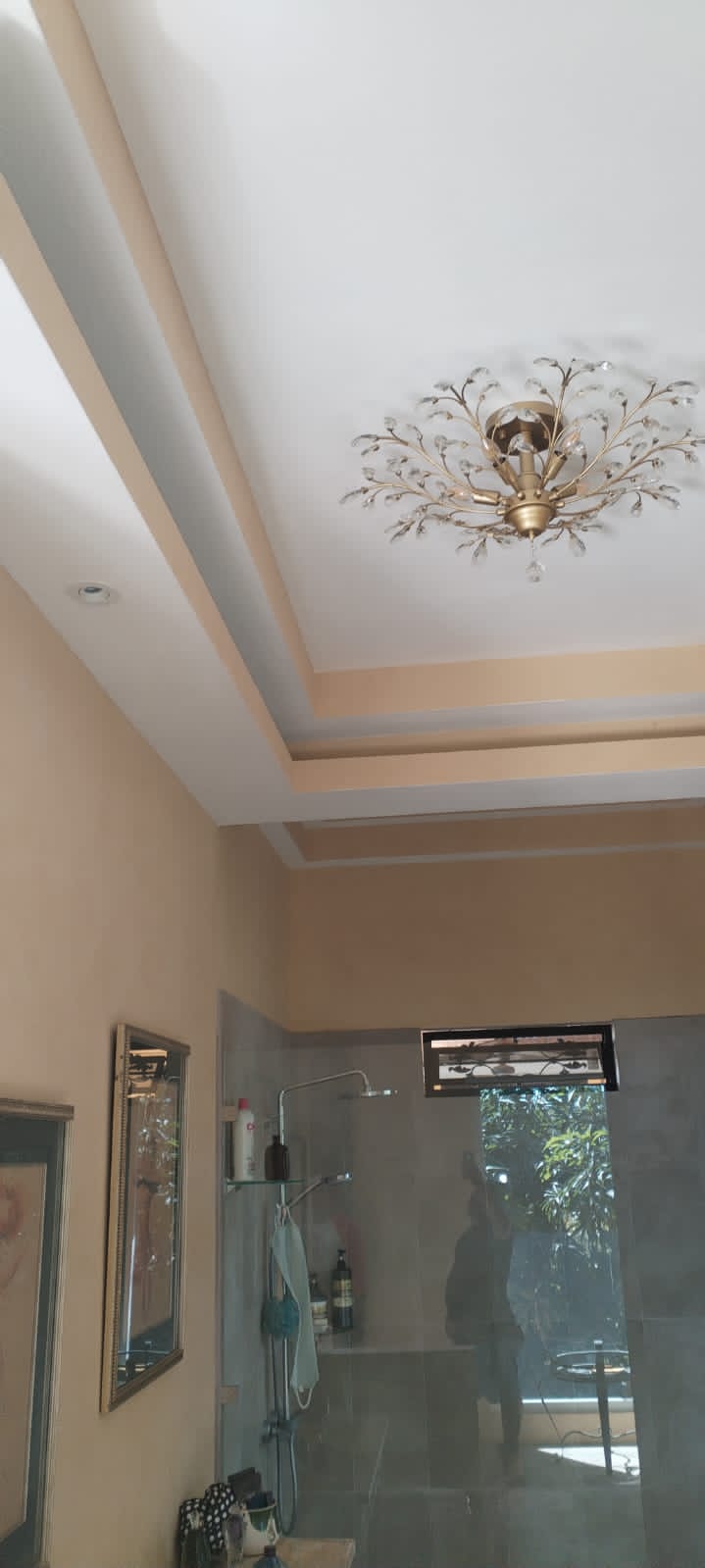 Vista de un baño con paredes en tonos beige, una lámpara de techo decorativa y una ducha con mampara de vidrio.