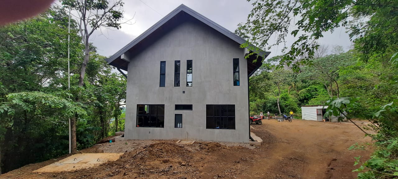 Se observa la fachada exterior de una casa de estilo moderno con paredes de concreto gris y marcos de ventanas negros rodeada de vegetación.
