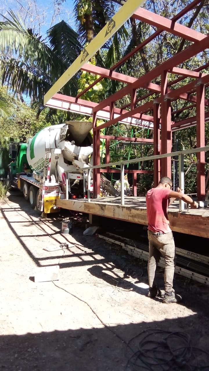 Un trabajador realiza labores de construcción en una estructura metálica mientras un camión mezclador de cemento está presente en el sitio.