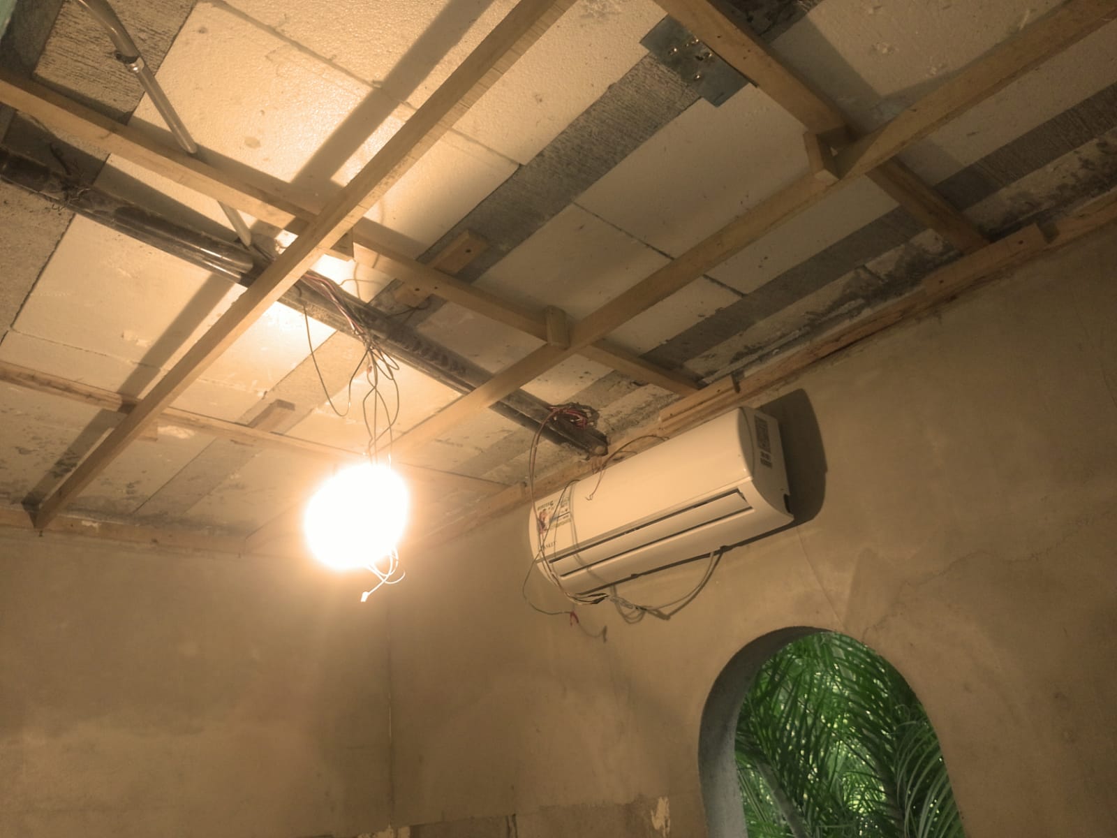 Se observa el interior de una habitación en construcción con una estructura de madera en el techo, una lámpara colgante y un aire acondicionado instalado.