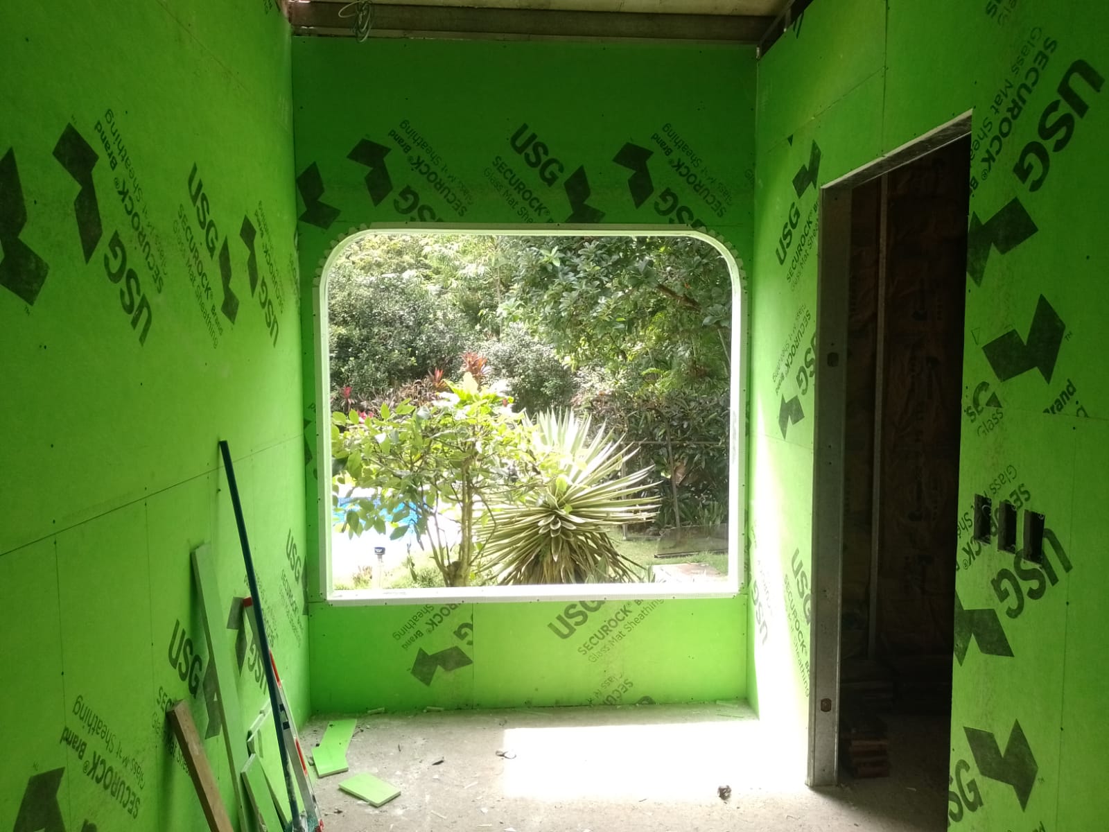 Una habitación en construcción con paredes revestidas de paneles verdes USG Securock y una ventana arqueada con vista al jardín.