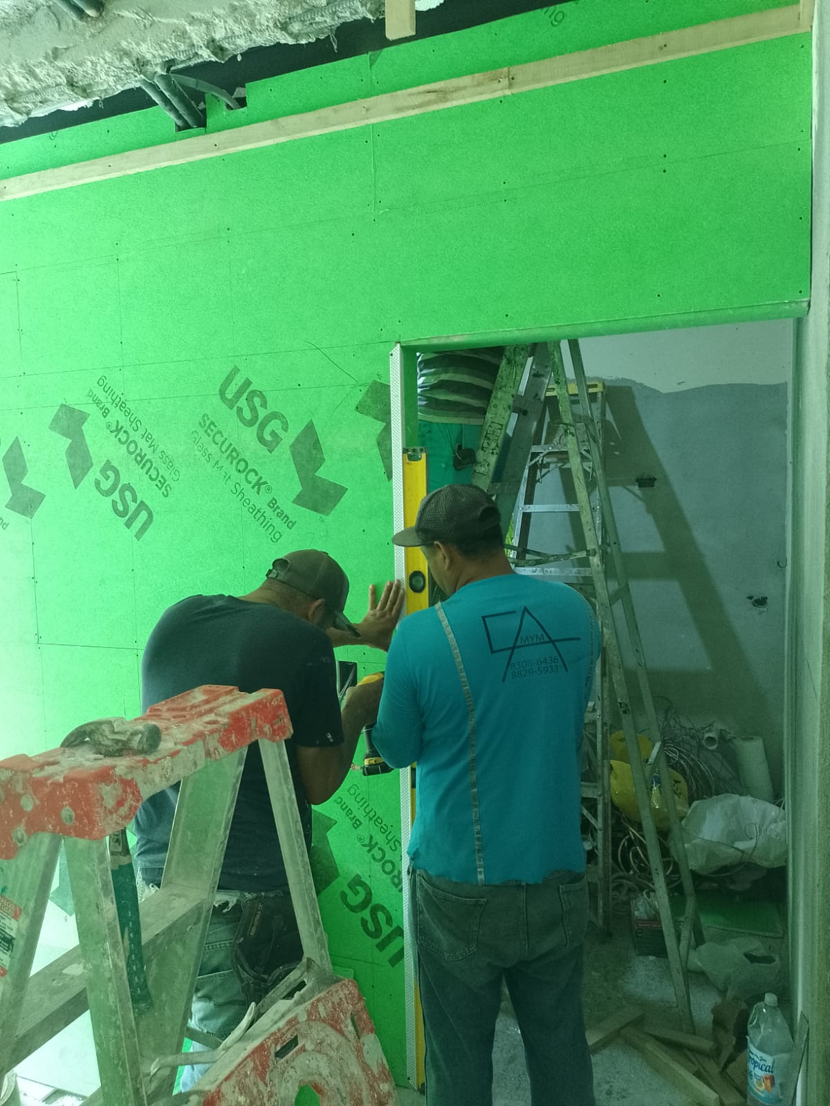 Dos trabajadores instalan paneles de yeso verde en una pared interior utilizando un nivel.