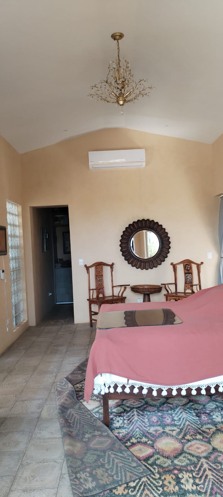 Vista interior de una habitación con paredes color crema, muebles de madera, un espejo circular y una cama con colcha rosada.