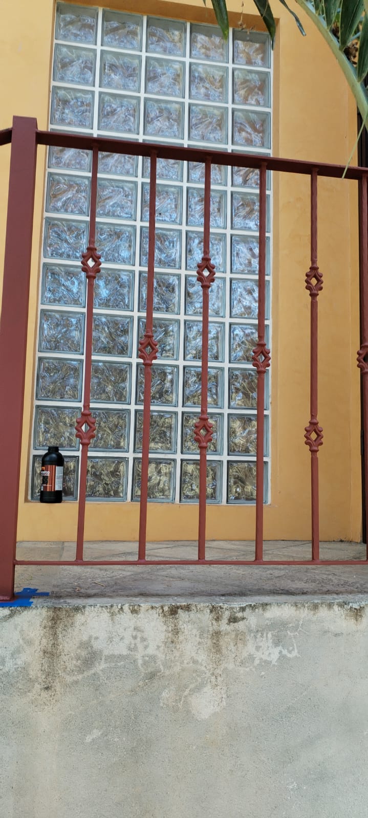Se observa una pared de color amarillo con una ventana de bloques de vidrio y una barandilla de metal rojizo en primer plano.