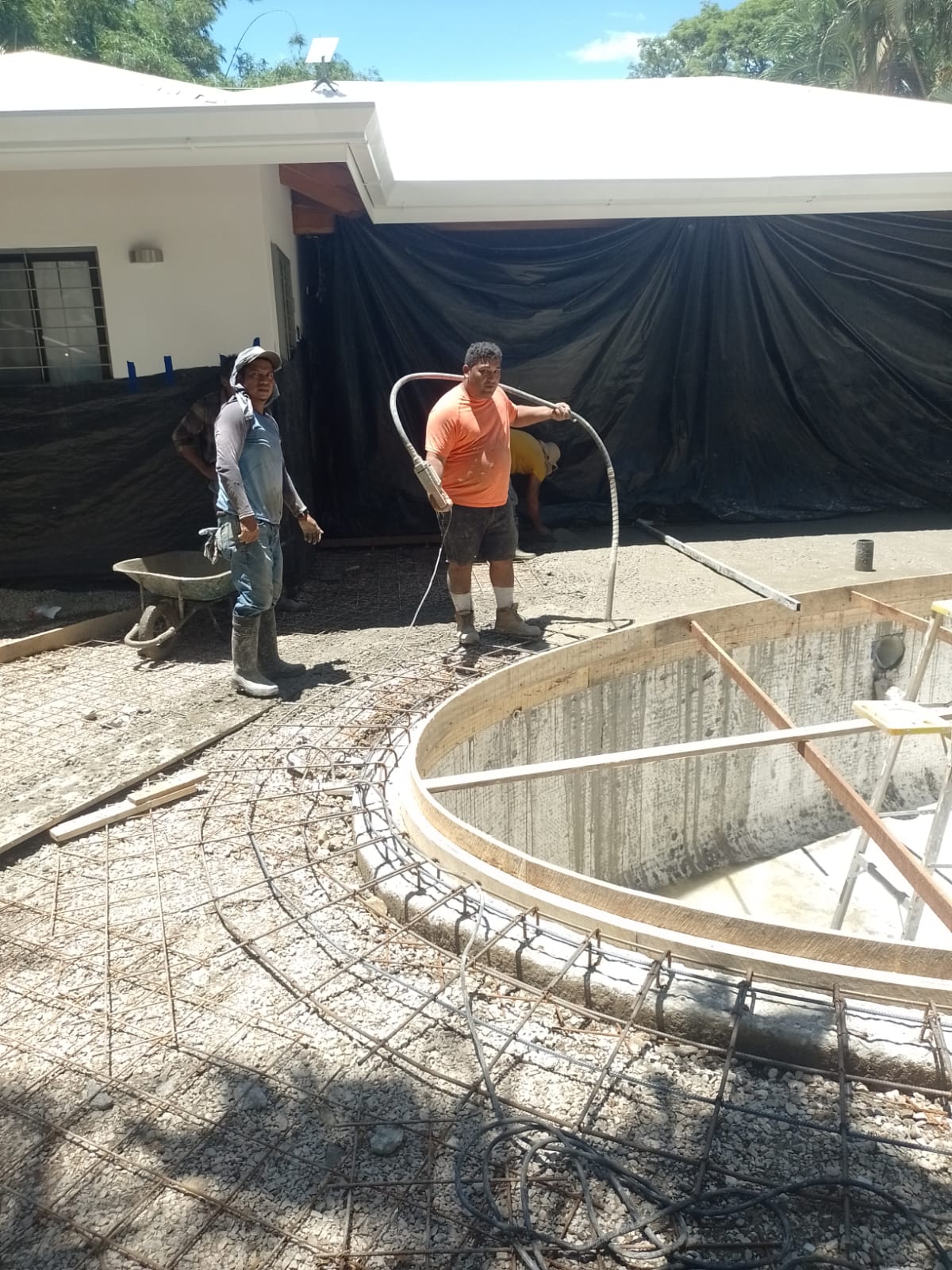 Dos trabajadores realizan labores de construcción y vertido de concreto alrededor de una estructura circular para una piscina.