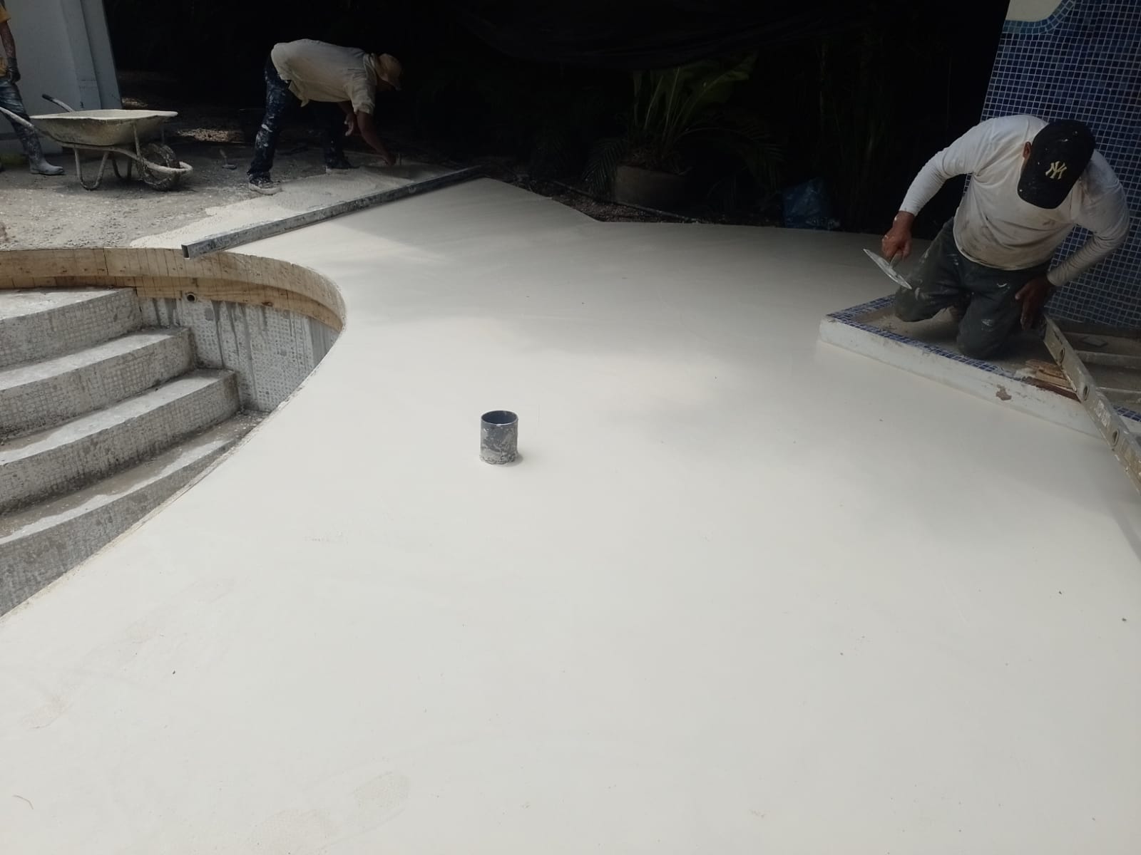 Dos trabajadores están aplicando y alisando cemento fresco alrededor de una piscina en construcción.