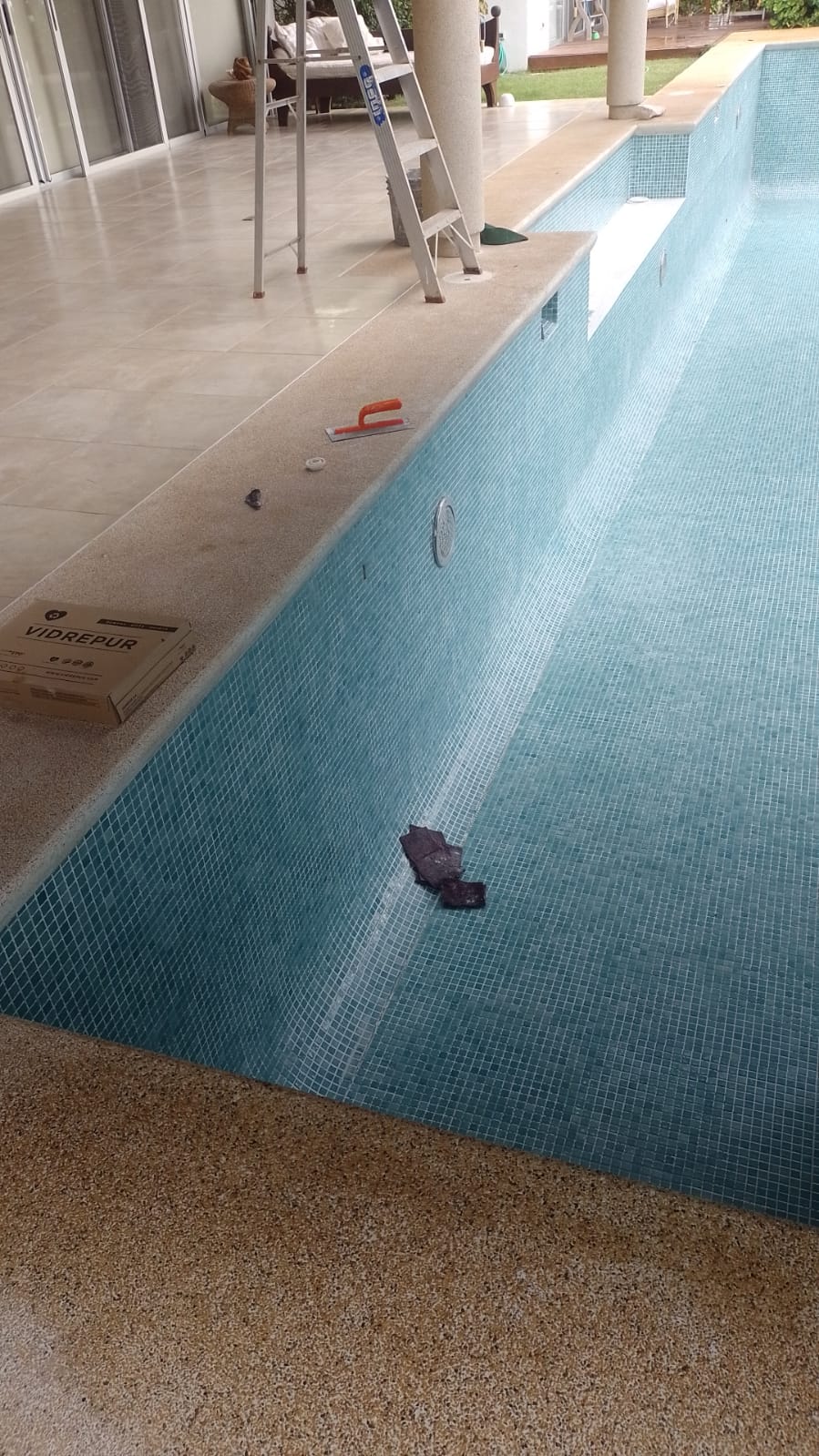 Se observa una piscina en proceso de instalación de azulejos con herramientas de trabajo y una caja de materiales Vidrepur a un lado.
