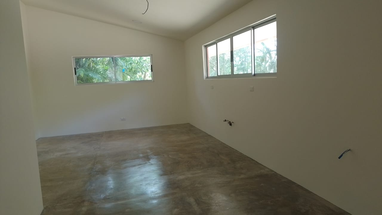 Una habitación interior vacía con paredes blancas, piso de cemento pulido y ventanas de aluminio.