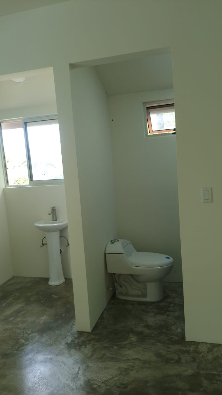 Se muestra un baño moderno con paredes blancas, piso de cemento pulido, un inodoro y un lavamanos de pedestal.