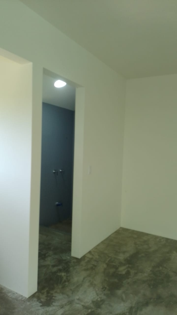 Se observa el interior de una habitación con paredes blancas y un piso de concreto pulido, con una entrada hacia un baño de pared oscura.